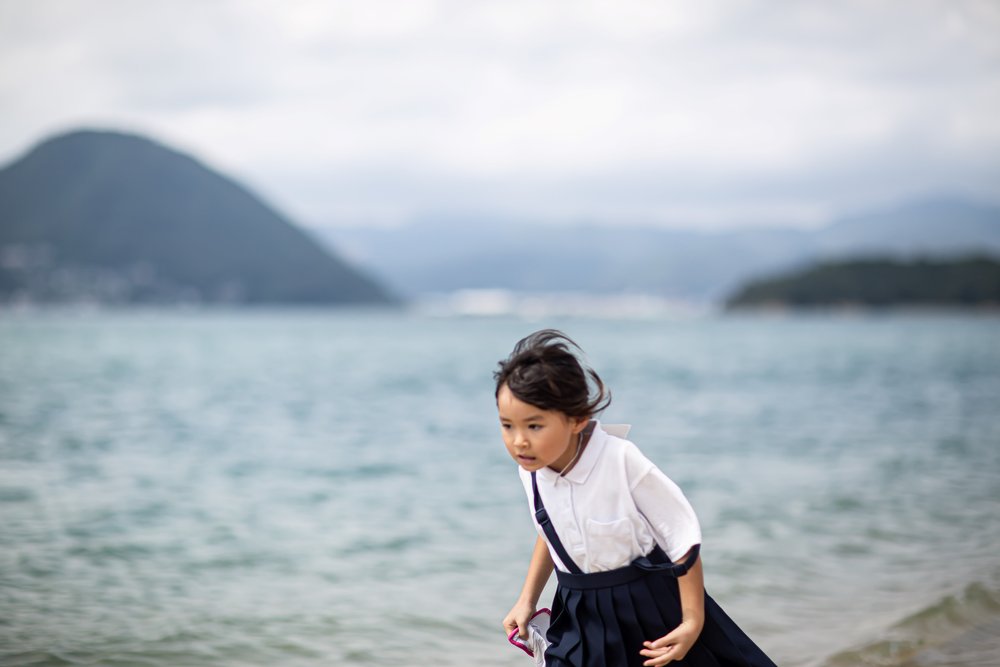 A Sagiura Elementary School student on the coast, mountains behind / 鷺浦小学校の児童、佐木島の海岸にて