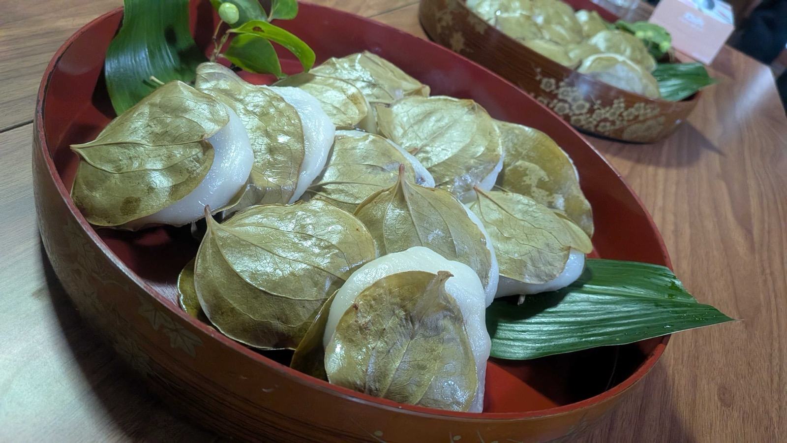 Close-up of the kashiwa-mochi, each cake wrapped in a sankirai leaf, with a leafy sprig of sankirai and its berries laid in the lacquered bowl. 柏餅の接写。一つ一つサンキライの葉で包まれ、実のついた小枝が添えられている。