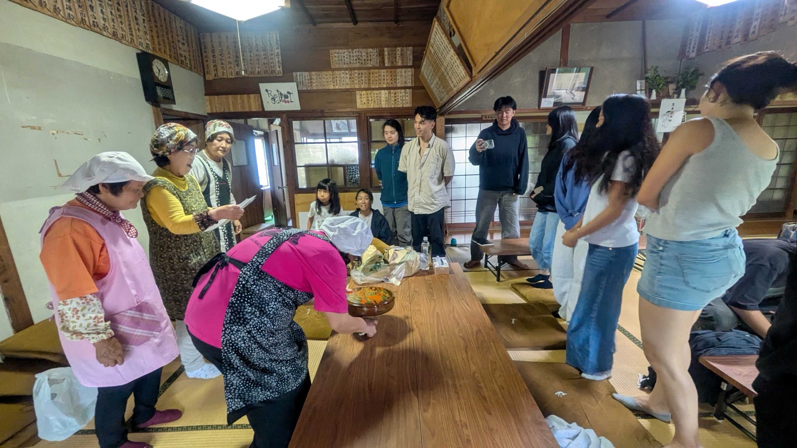 Island grandmothers at Kakumeisha; one reads the welcome letter aloud to the cohort, with the bowl of kashiwa-mochi on the table before them. 格明舎にて、島のおばあちゃんたちが歓迎の手紙を読み上げる。手前には柏餅。