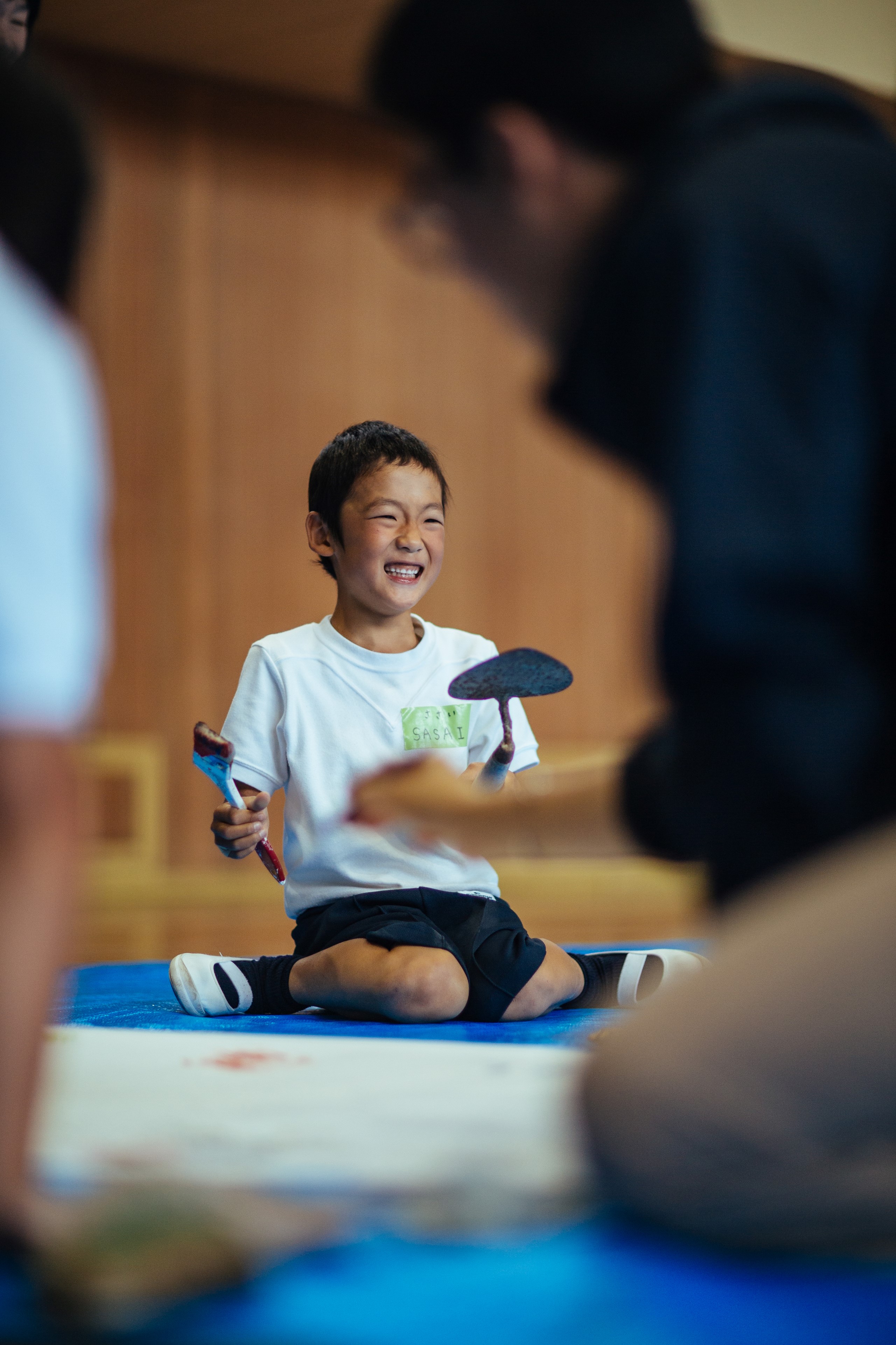 A laughing schoolchild from Sagiura Elementary sitting with the cohort, holding a hand-carved stamp during the ceremony printmaking. 鷺浦小学校の児童が、儀式の版づくりのなかで手彫りの版を手に、学生たちと並んで笑っている。