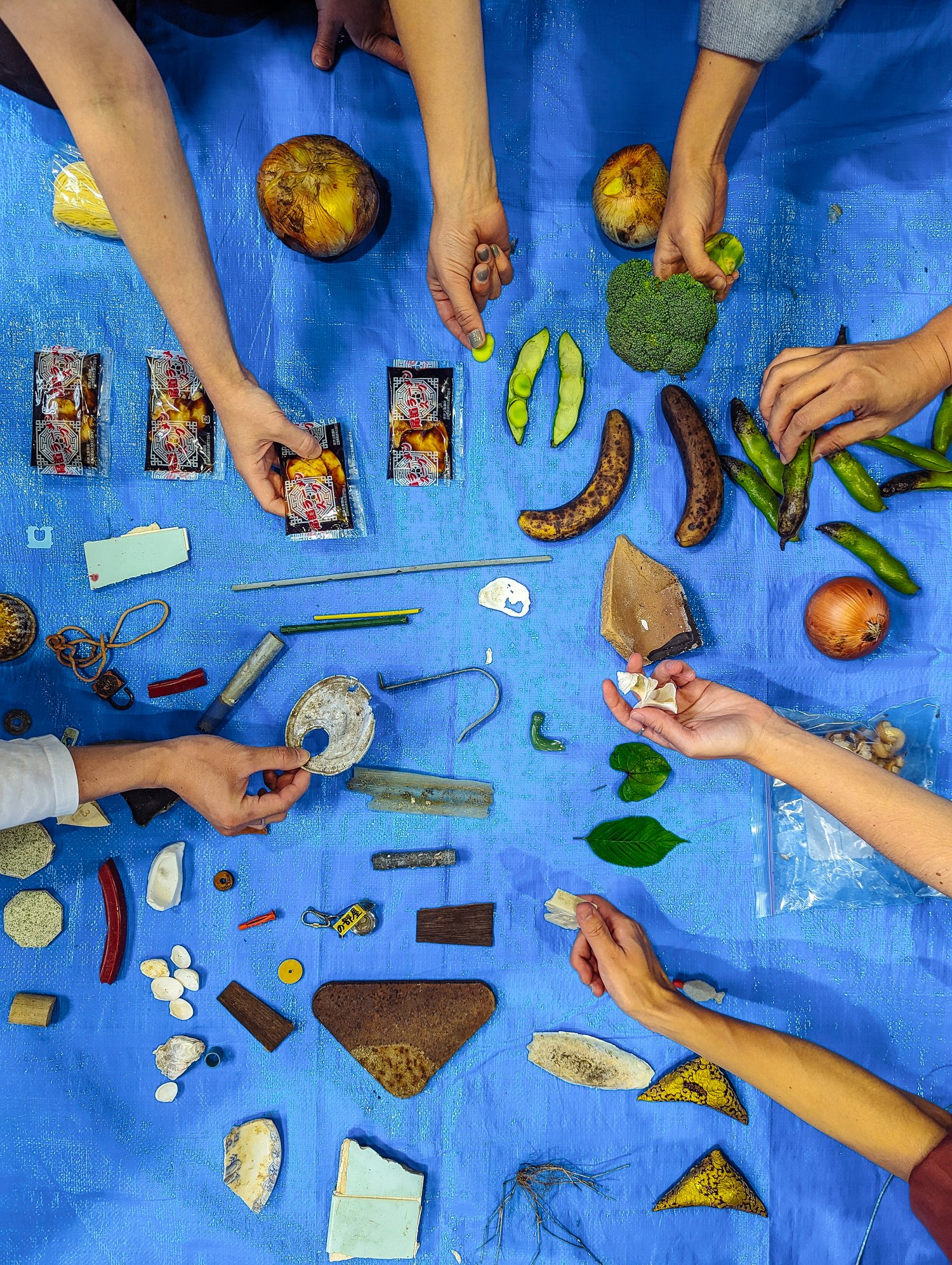 Hands sorting and cataloging found materials on a blue tarp: salvaged fragments, hardware, and produce laid out and examined one piece at a time. 青いシートの上で、見つけた素材を一つずつ並べて調べ、分類する手元。再生材の断片、金物、農作物が広げられている。