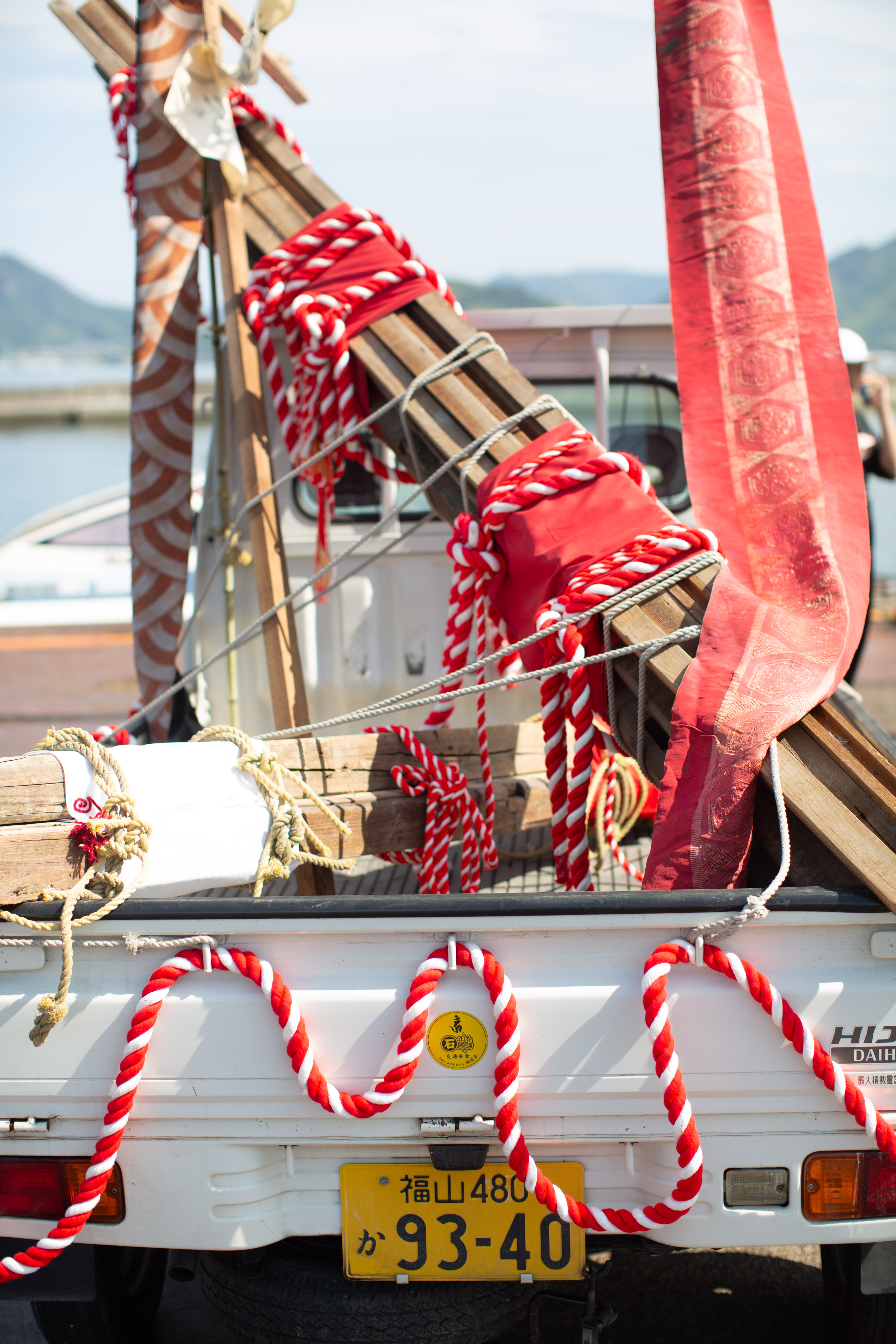 The deconstruction-ceremony regalia loaded on a light truck at the harbor: red-and-white braided rope, fish-scale-patterned cloth, and bound salvaged timber. 港で軽トラックに積まれた解体の儀式の祭具。紅白の組み紐、鱗文様の布、束ねられた再生木材。