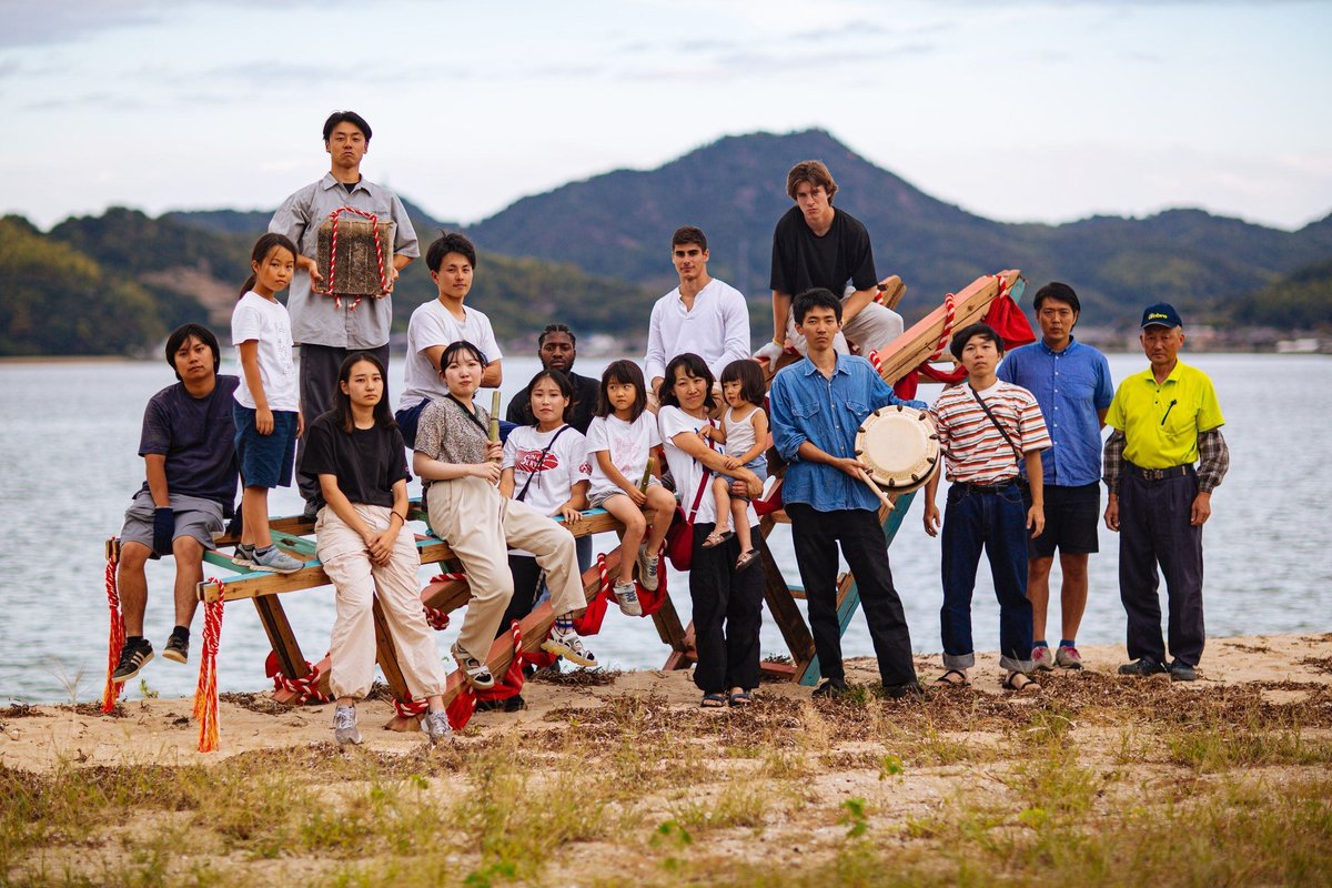 The September 2023 cohort with island residents and an elder on the Sagishima shore, holding the deconstruction-ceremony regalia: red-and-white rope, a drum, and decorated salvaged timber. 2023年9月のコホートが、島の住民や年配の方とともに佐木島の浜辺に並び、解体の儀式の祭具（紅白の縄、太鼓、装飾された再生木材）を手にしている。