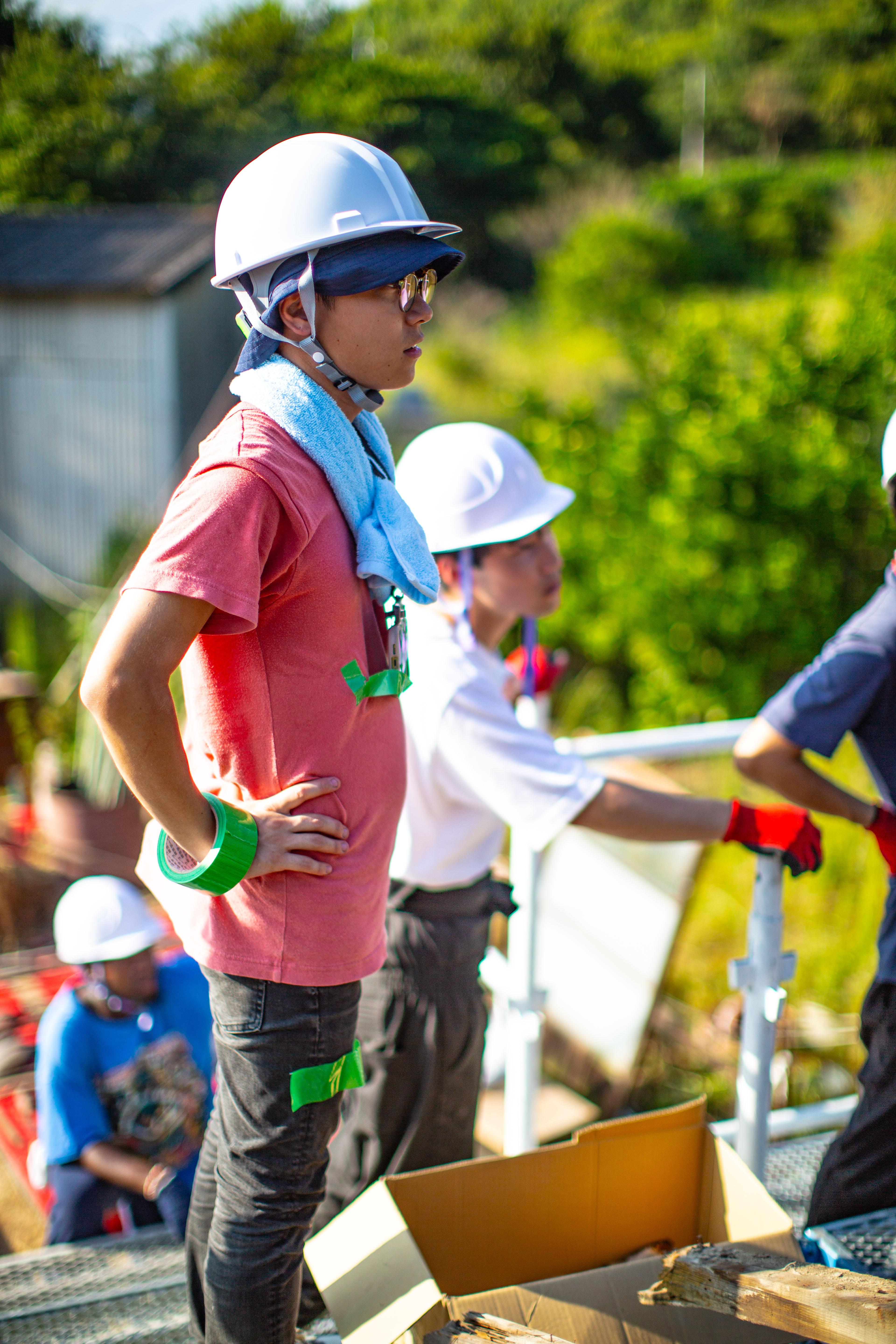 A student in a hard hat at the deconstruction worksite under bright sun, a towel around the neck, with other masked-and-helmeted workers handling material. 強い日差しの下、解体の現場でヘルメットをかぶり首にタオルを巻いた学生。ヘルメットとマスク姿の作業者たちが資材を扱っている。