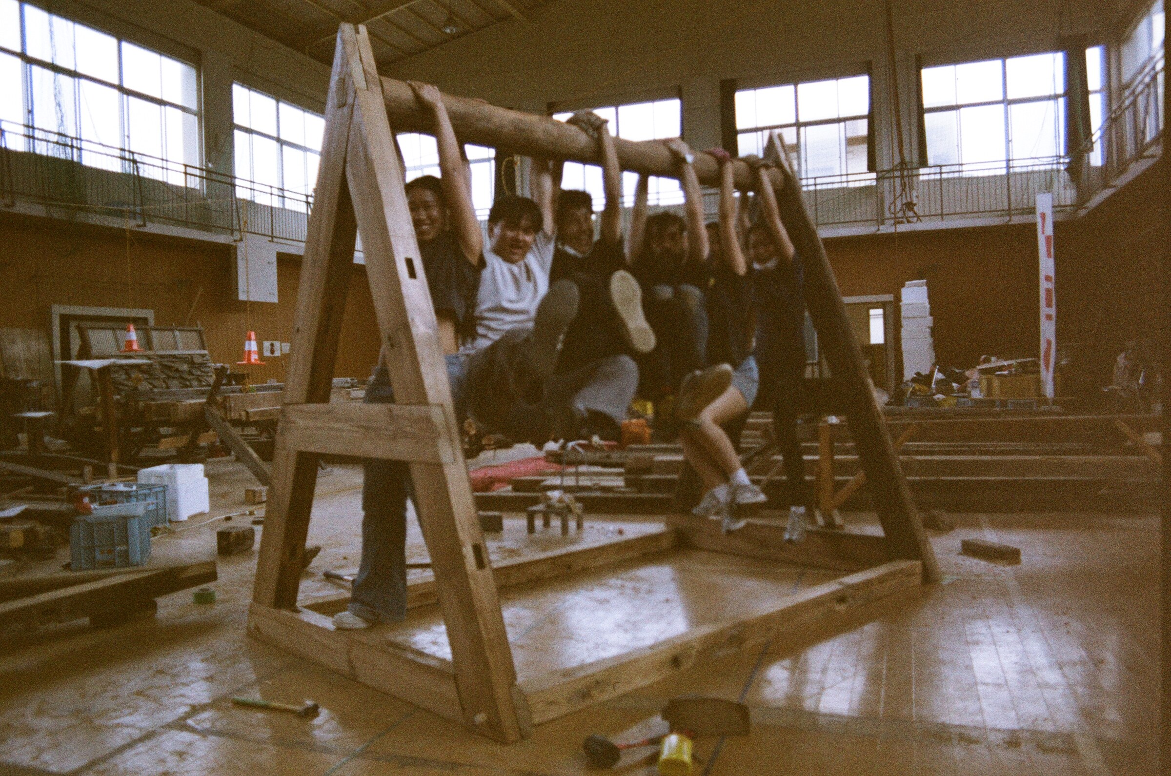 Members of the Studio A1 cohort hanging in a row from the ridge log of a full-size timber A-frame raised on the floor of the old school gymnasium, with salvaged lumber, sawhorses, and a sill frame laid out around them. 旧校舎の体育館の床に立ち上げられた原寸大の木造の合掌の棟木に、スタジオ A1 のコホートが一列にぶら下がっている。まわりには再生材の木材、木挽き台、土台の枠が並べられている。