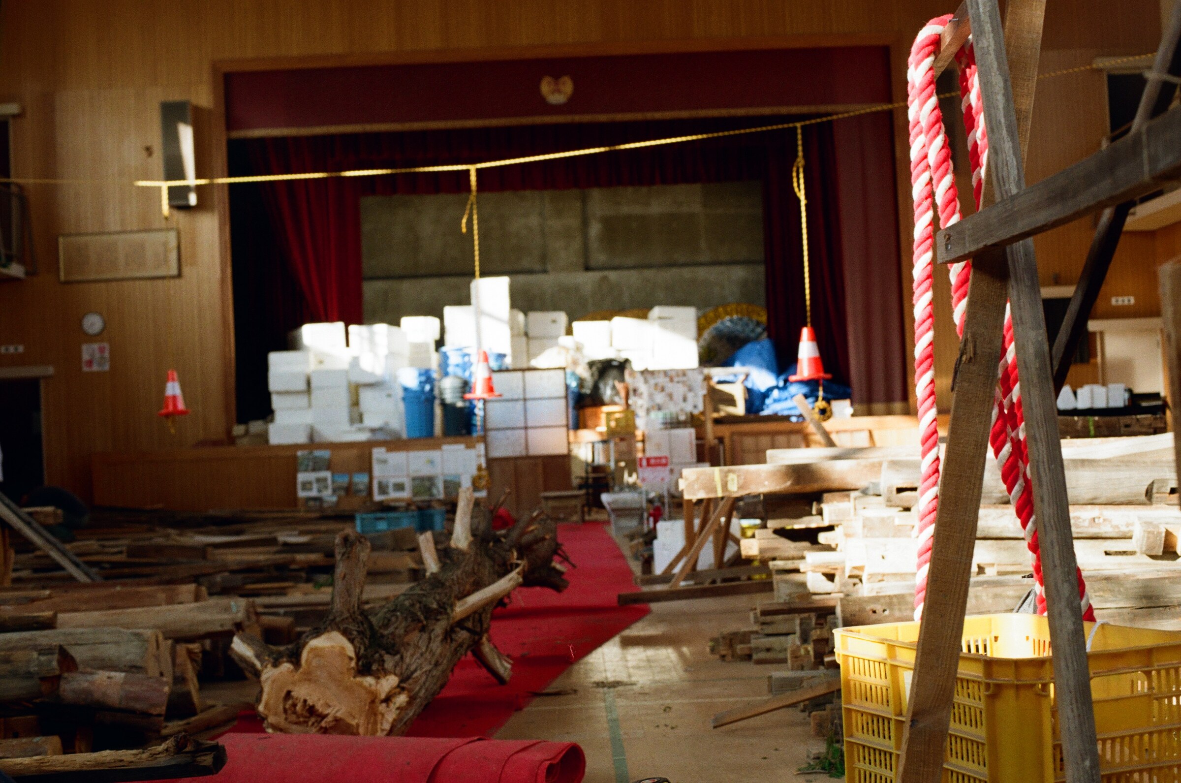 The old school auditorium turned salvage yard: recovered timber laid out in sorted rows across the floor, a gnarled root mass in the foreground, stacked white crates filling the curtained stage, and red-and-white braided rope hung from a raised timber frame. 旧校舎の講堂がサルベージの置き場となっている。回収された木材が床一面に仕分けて並べられ、手前には根株が横たわる。緞帳の上がった舞台には白い箱が積み上げられ、立ち上げられた木の骨組みからは紅白の綱が下がっている。