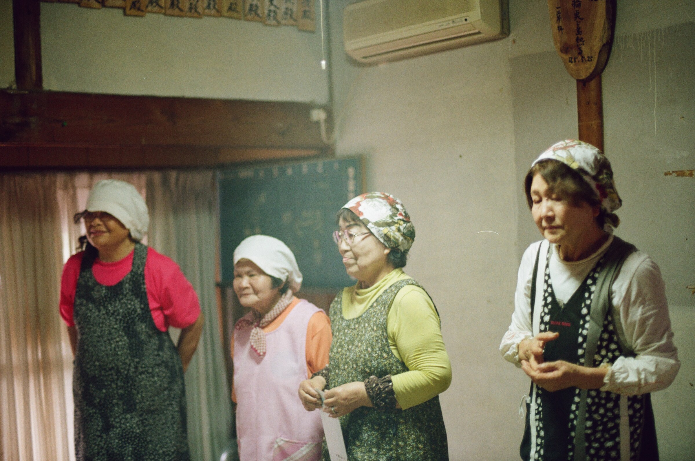 Four island women in work aprons and headscarves standing in a community room, one holding a printed sheet, with a chalkboard, a dedication paddle, and rows of wooden donor plaques on the wall behind them. 集会所の一室に並ぶ、仕事着の割烹着と姉さんかぶりの島の女性四人。一人は印刷された紙を手にしている。背後の壁には黒板と奉納の櫂、そして寄進者名を記した木札が並んでいる。