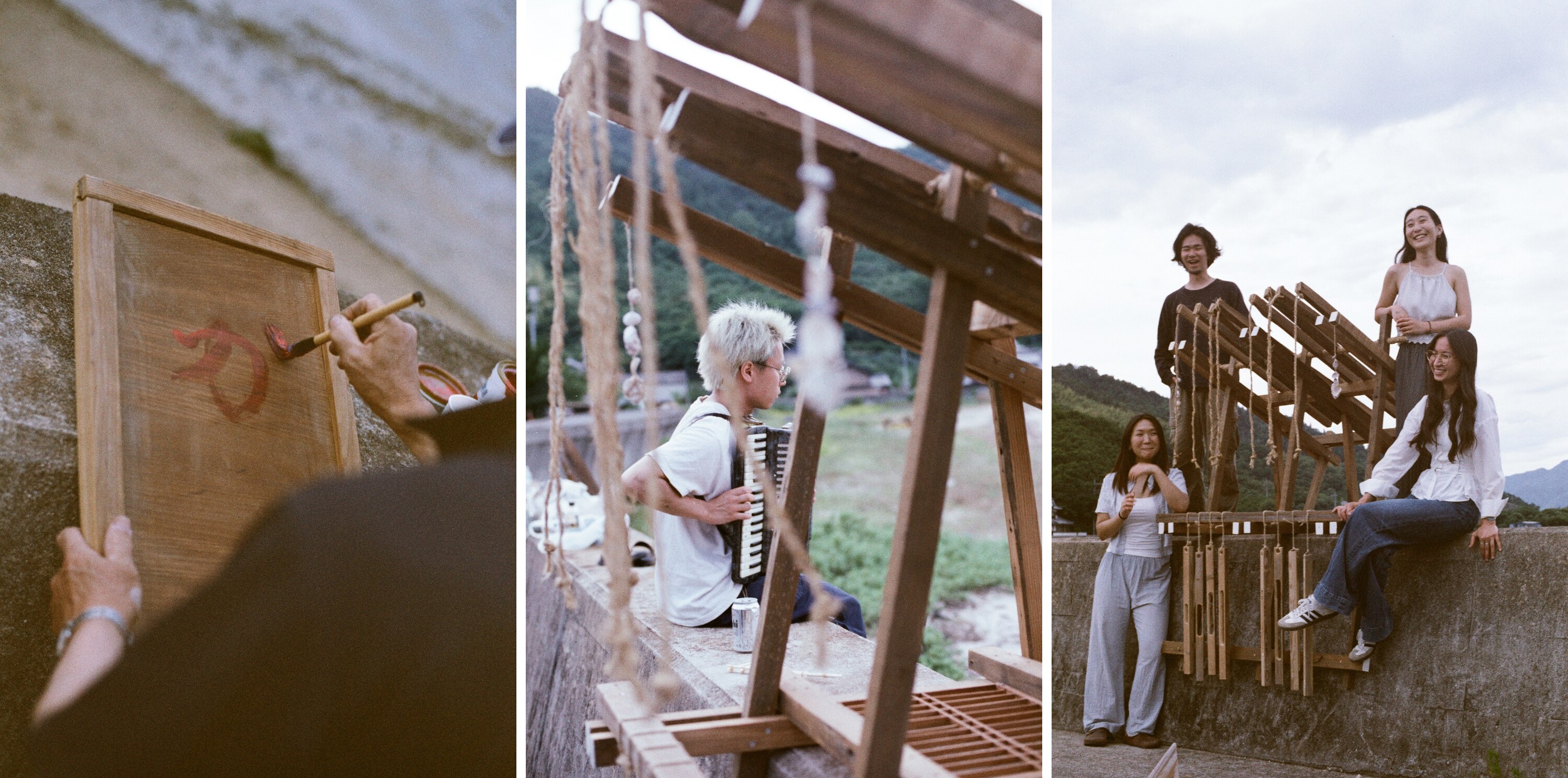 A three-panel sequence made on the seawall at dusk. Left: hands brushing a red mark onto a framed wooden panel. Centre: a studio member seated on the wall playing an accordion, seen through the raking beams and hanging ropes of the timber rack. Right: four studio members with the finished rack they built, wooden slats and cords hung along its frame. 夕暮れの護岸で撮られた三枚組。左は、木枠のついた板に赤い印を刷毛で描く手元。中央は、木の架構の斜めの梁と垂れた縄越しに見える、護岸に腰かけてアコーディオンを弾くスタジオの一人。右は、自分たちで組み上げた架構とともに立つスタジオの四人。枠には木の板と紐が吊るされている。