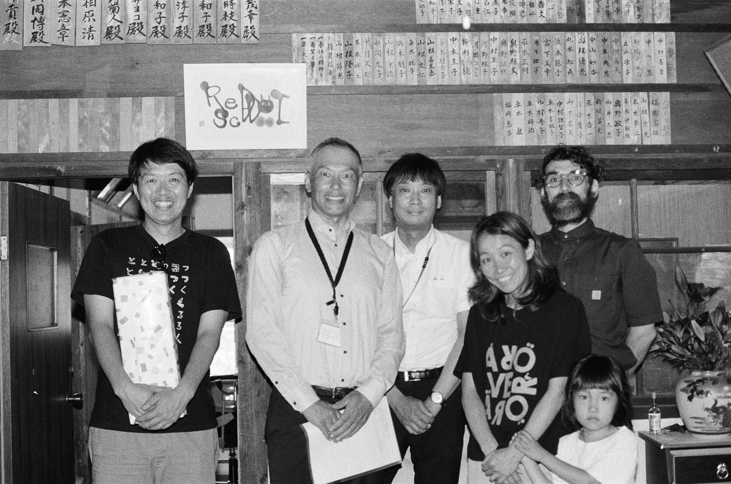 Black-and-white photograph of five adults and a child standing together in a traditional timber-framed room, one holding a wrapped gift, beneath a wall lined with rows of vertical wooden dedication plaques; a hand-lettered "Red Dot School" sign hangs framed on the post behind them. 白黒写真。伝統的な木造の部屋に、大人五人と子ども一人が並んで立っている。一人は包装された贈り物を手にしている。壁一面には奉納者名を記した木札が並び、背後の柱には手書きの「Red Dot School」の額が掛かっている。