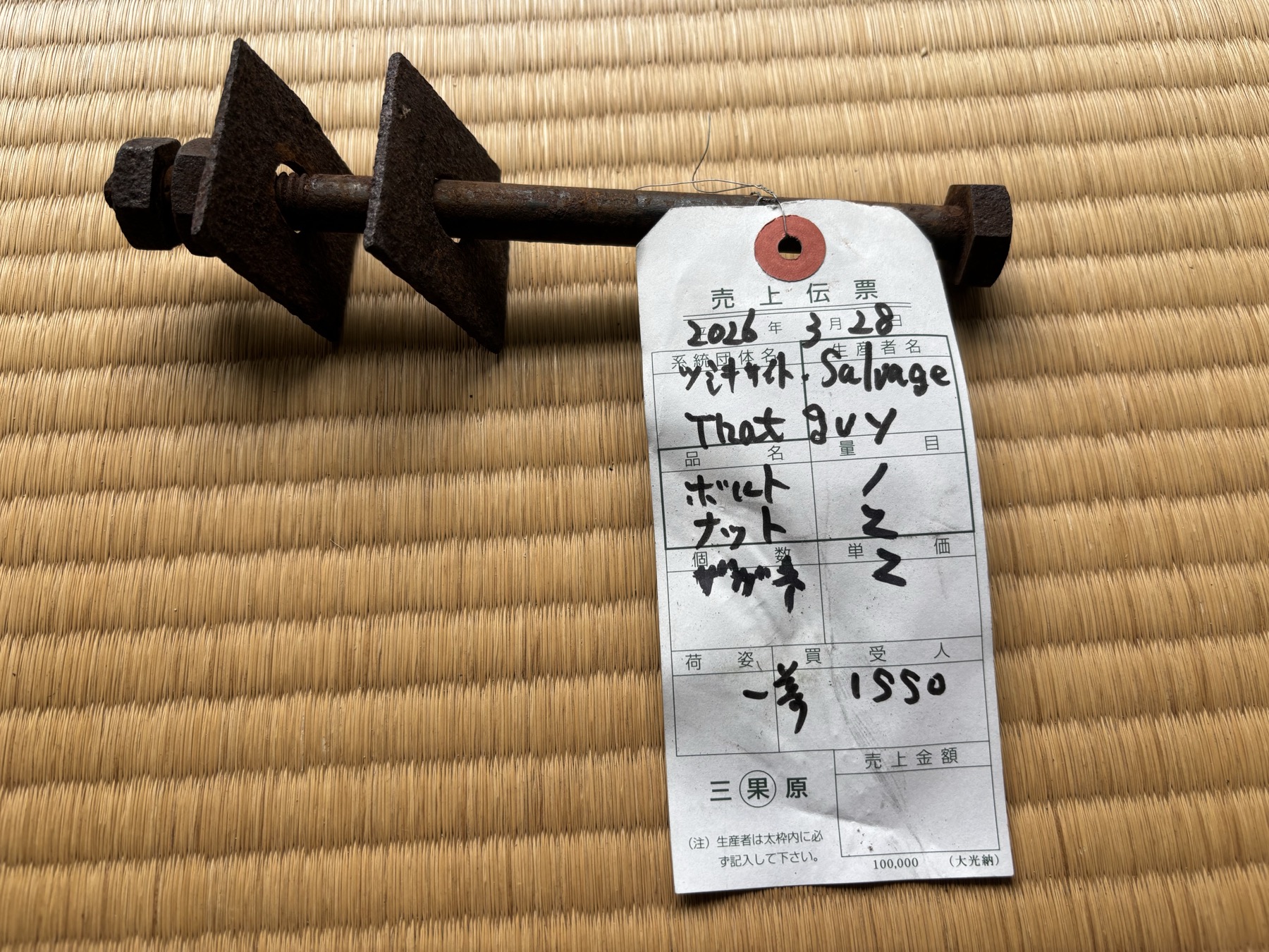 A salvage tag on a repurposed sales-slip, tied to an iron tie-bolt and its two diamond washer-plates from the Nakamura house; hand-marked "ナカムラ Salvage / That guy," 2026-03-28 / 中村家から救い出した鉄の通しボルトと二枚の菱形座金に結ばれた、売上伝票を転用した荷札。「ナカムラ Salvage / That guy」と手書きされている(2026年3月28日)。