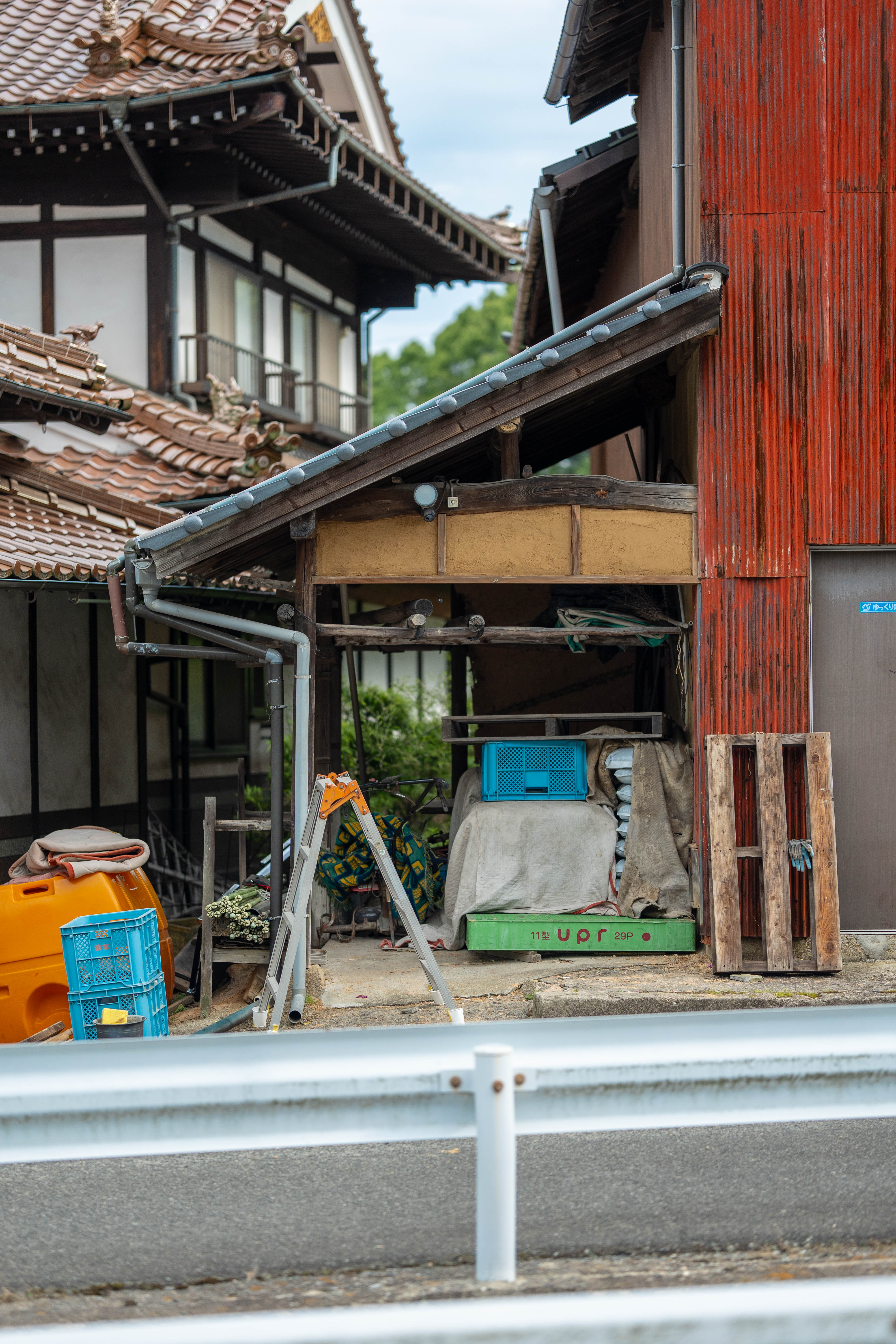 A street view of traditional island houses with an open salvage-storage bay: roof tiles, timber, and blue crates gathered under the eaves. 島の伝統的な家並みと、軒下に屋根瓦・木材・青いコンテナを集めた、開かれたサルベージ置き場。