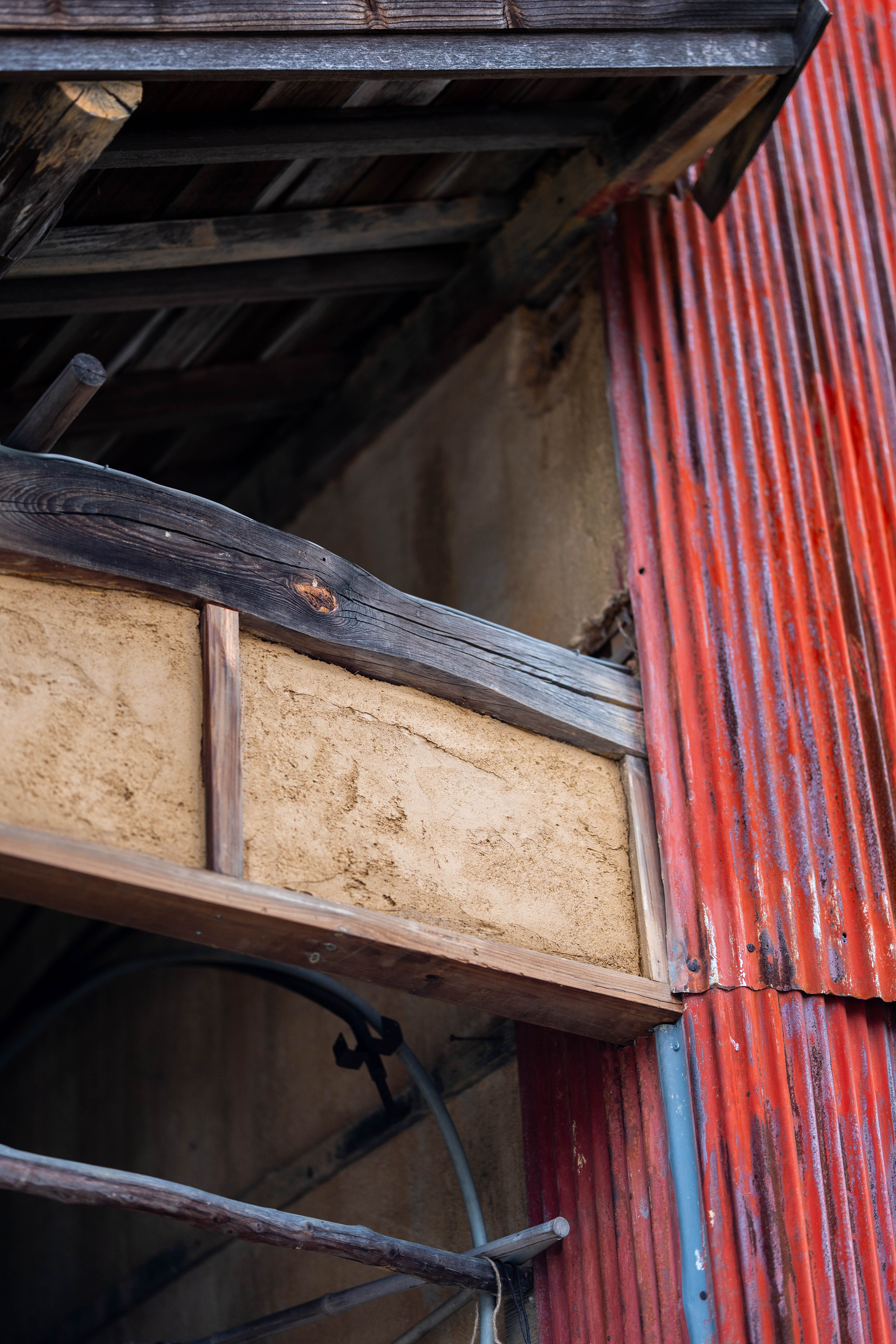 An earthen wall panel under the eaves beside a red corrugated-metal wall, the timber frame and clay infill visible. 軒下の土壁のパネルと、その隣の赤いトタンの壁。木の骨組みと土の充填がよく見える。